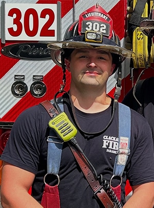 A firefighter in uniform and helmet stands in front of a fire truck with the number 302 visible on both his helmet and the truck.